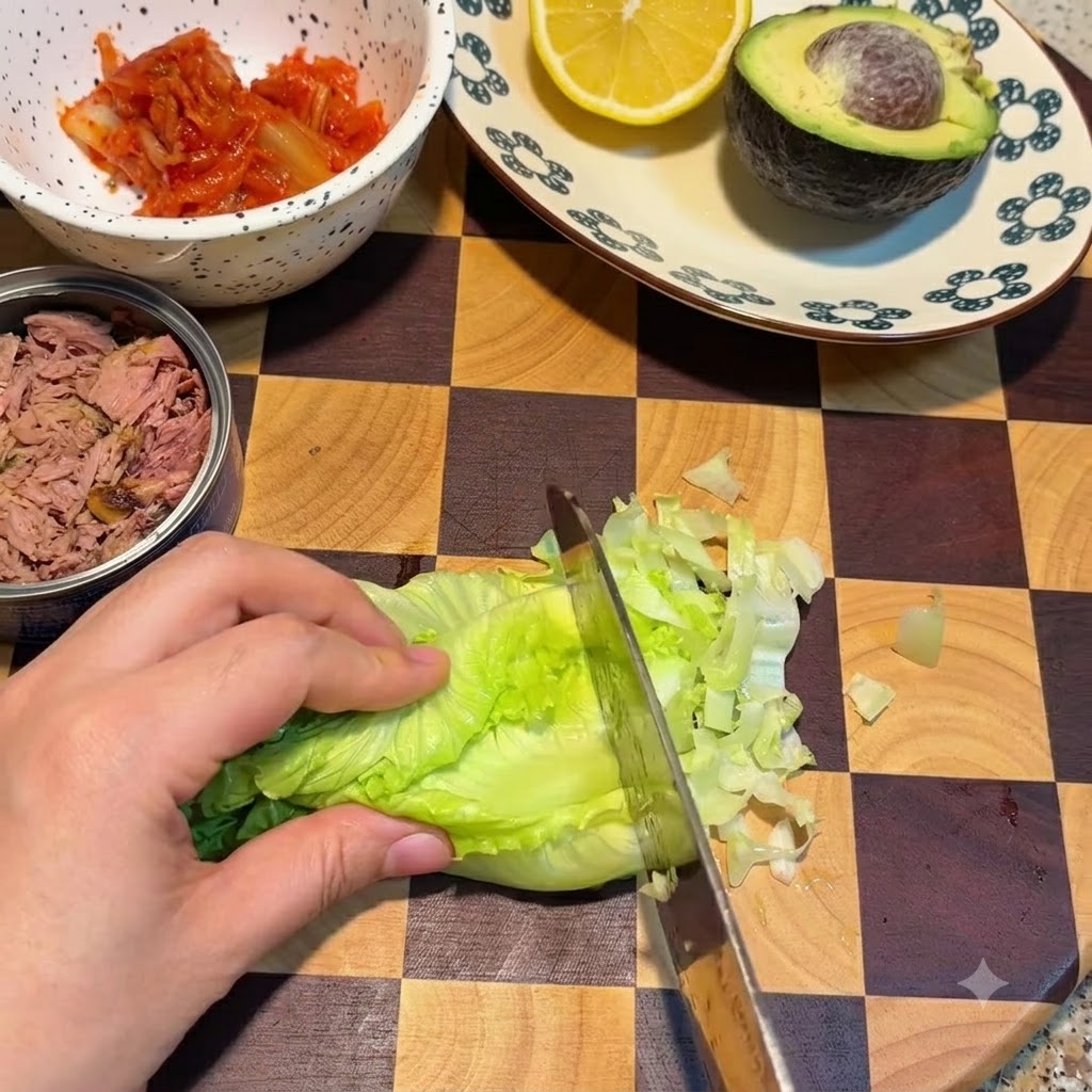 Un couteau coupant des feuilles de laitue verte fraîche sur une planche à découper à côté dun bol de kimchi et dune boîte de thon.