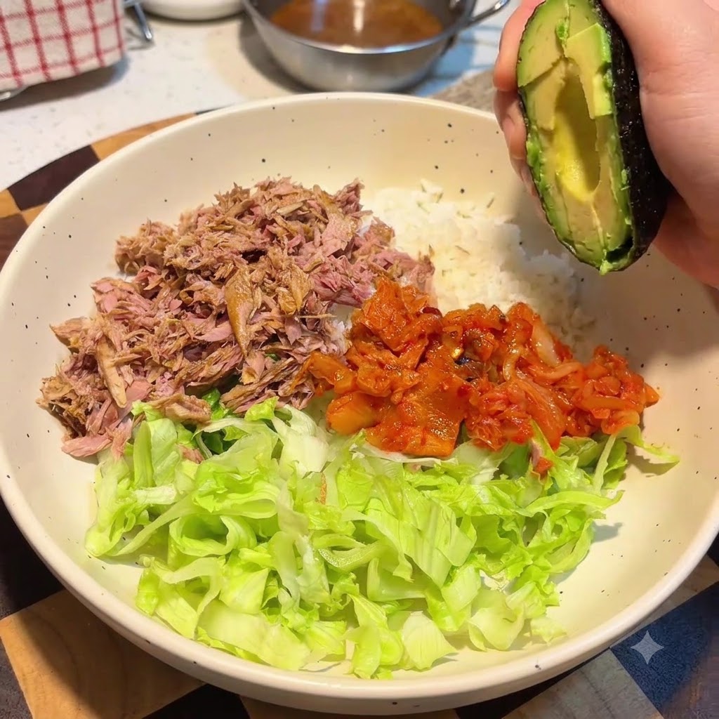 Une main tenant une moitié davocat au-dessus dun grand bol contenant du riz, de la laitue, du thon et du kimchi.