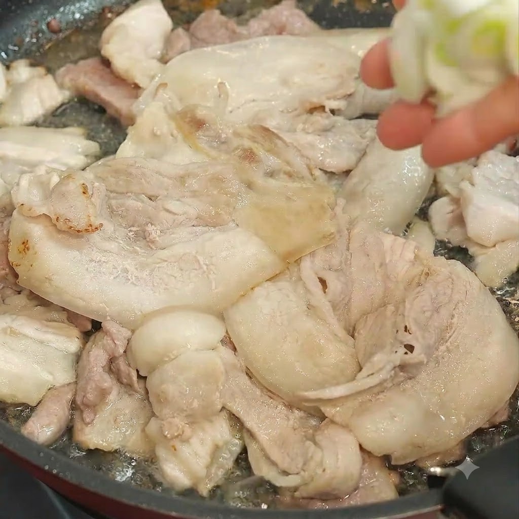 Une main jetant des oignons verts hachés dans une poêle remplie de tranches de porc en train de frire.