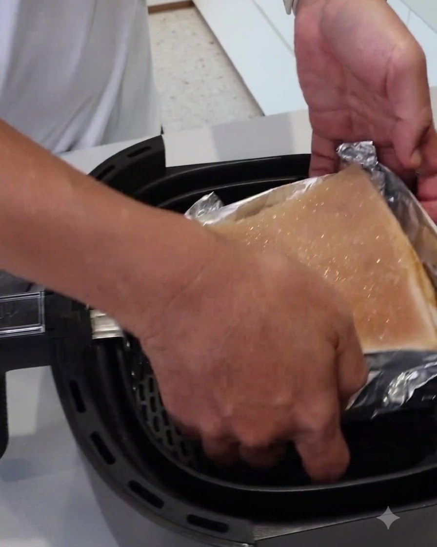 Une paire de mains abaissant un bloc de viande enveloppé de papier aluminium dans le panier dun air fryer.