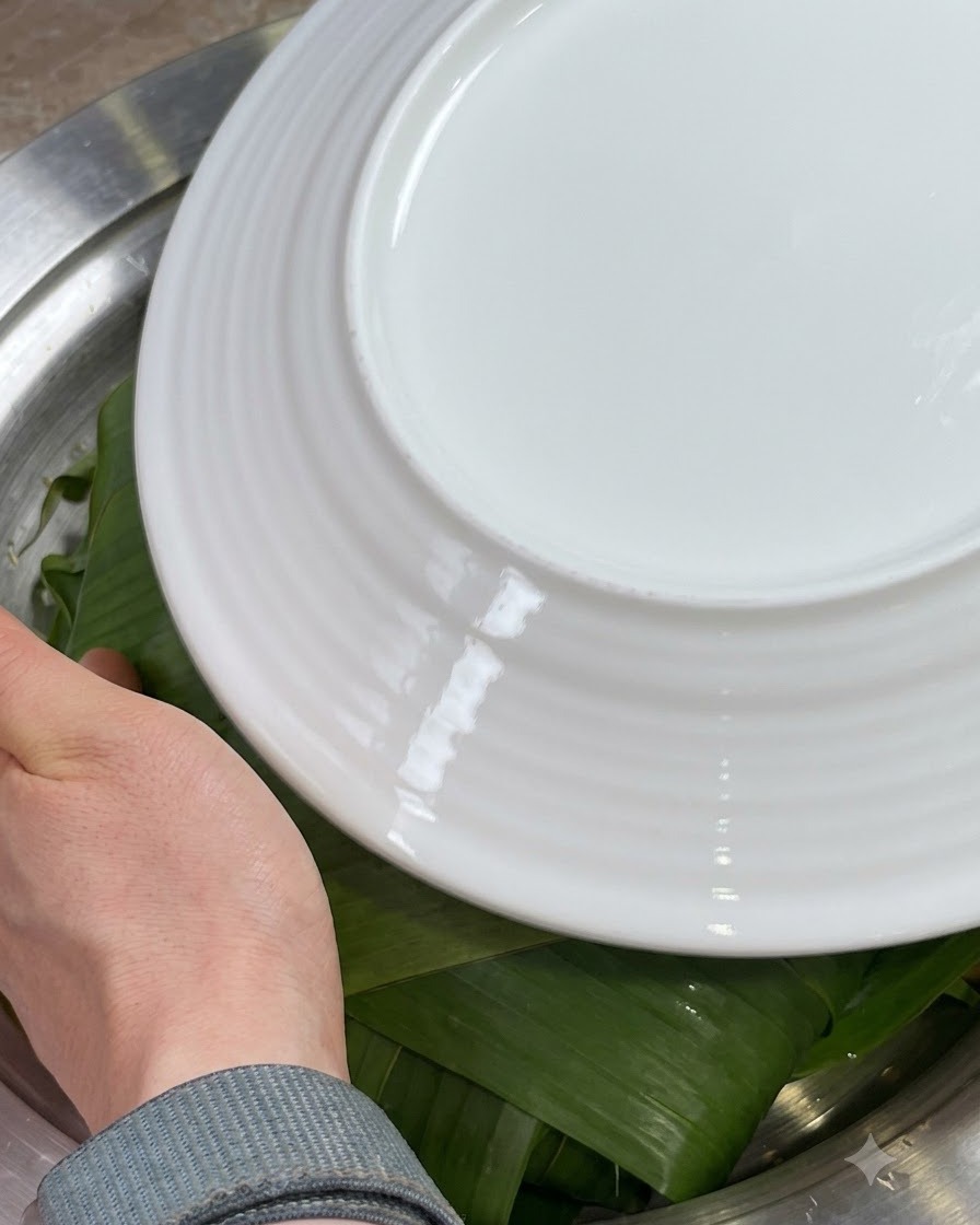 Mains repliant les bords des feuilles de Zong vertes vers lintérieur par-dessus les ingrédients, couverts par une assiette blanche.
