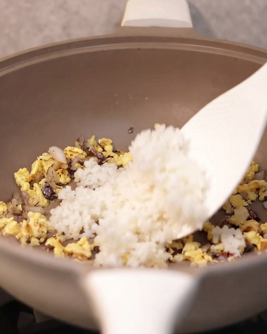 Une spatule ajoutant du riz blanc cuit dans la poêle avec les ingrédients assaisonnés.