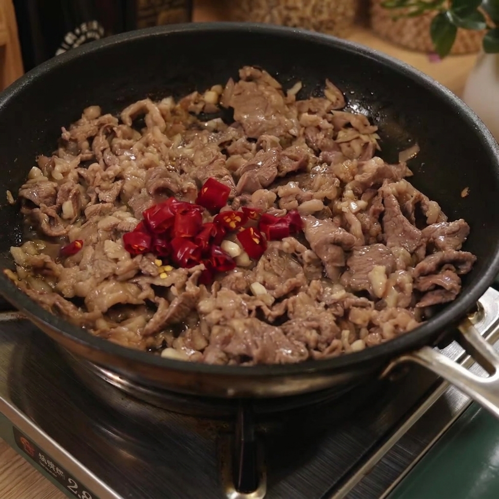 Des spaghettis cuits ajoutés dans une poêle avec du bœuf et des piments rouges tranchés.