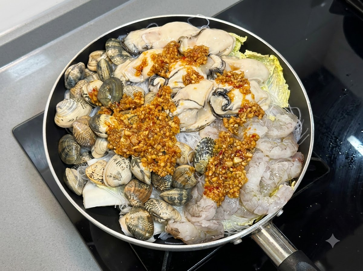 Sauce à lail et au piment cuite généreusement versée sur les couches de fruits de mer crus et de nouilles dans une poêle.