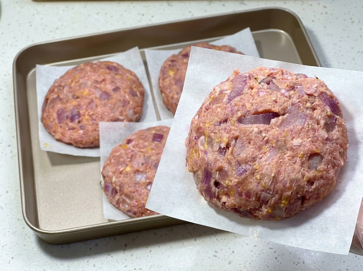 Un steak haché cru fraîchement formé sur du papier sulfurisé, avec dautres steaks disposés sur un plateau en arrière-plan.