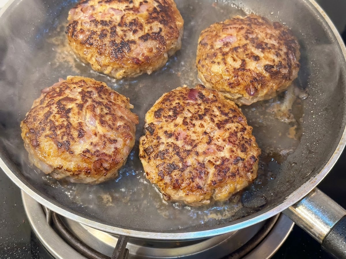 Quatre steaks hachés épais cuisant dans une poêle avec de leau et de la vapeur séchappant sous un couvercle.