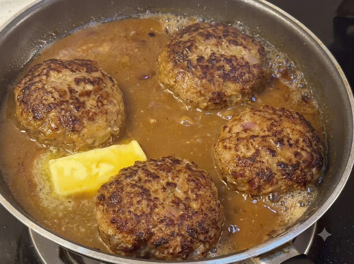 Des steaks hachés mijotant dans une sauce brune épaisse aux côtés dun morceau de beurre en train de fondre.