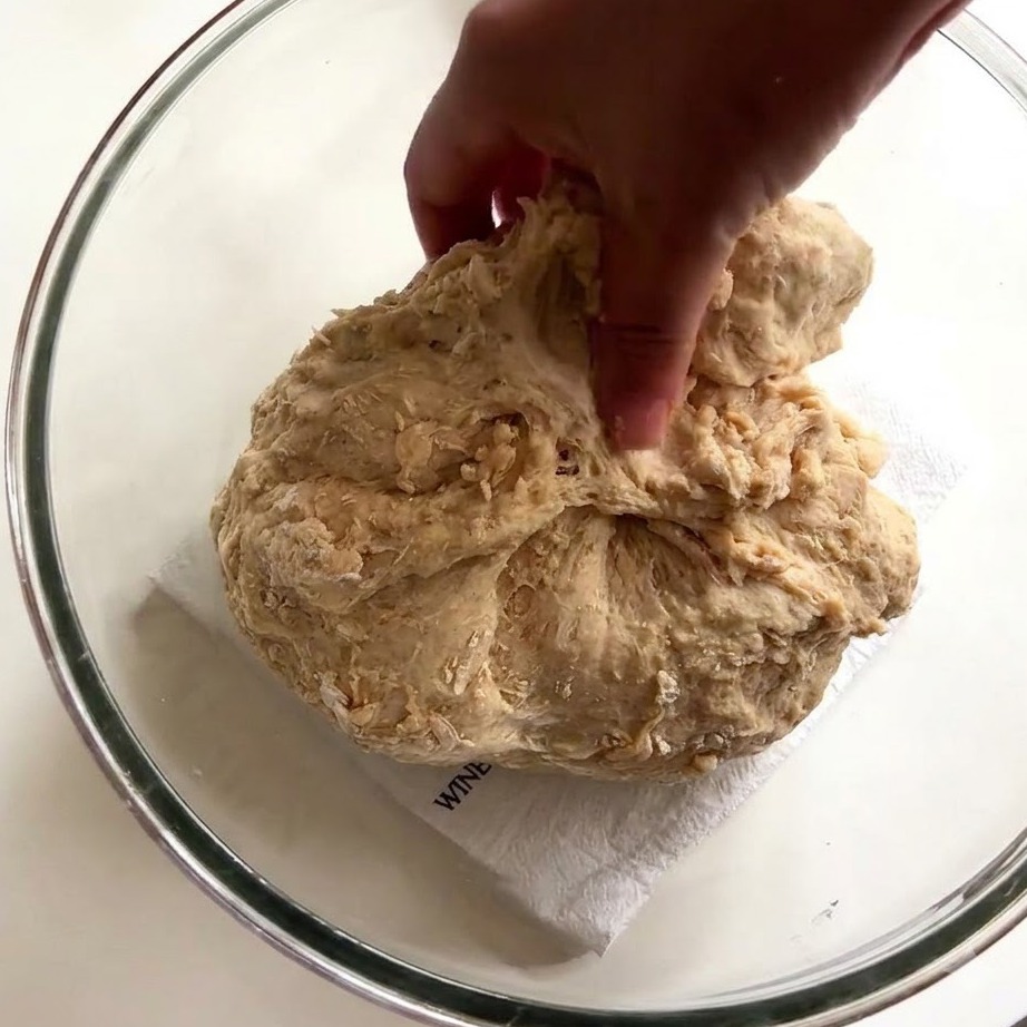 La main d une personne utilisant des doigts humides pour étirer et replier une pâte à pain collante à l intérieur d un bol en verre.