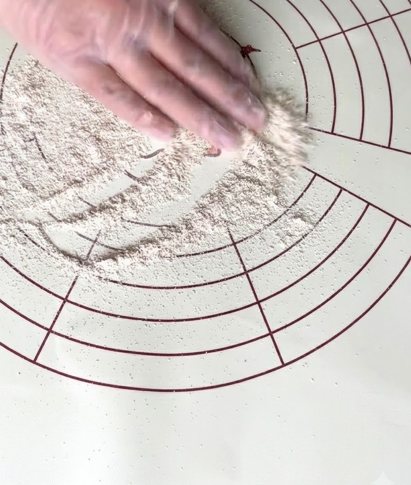 Une main gantée répartissant de la farine complète sur un tapis de pétrissage circulaire en silicone.