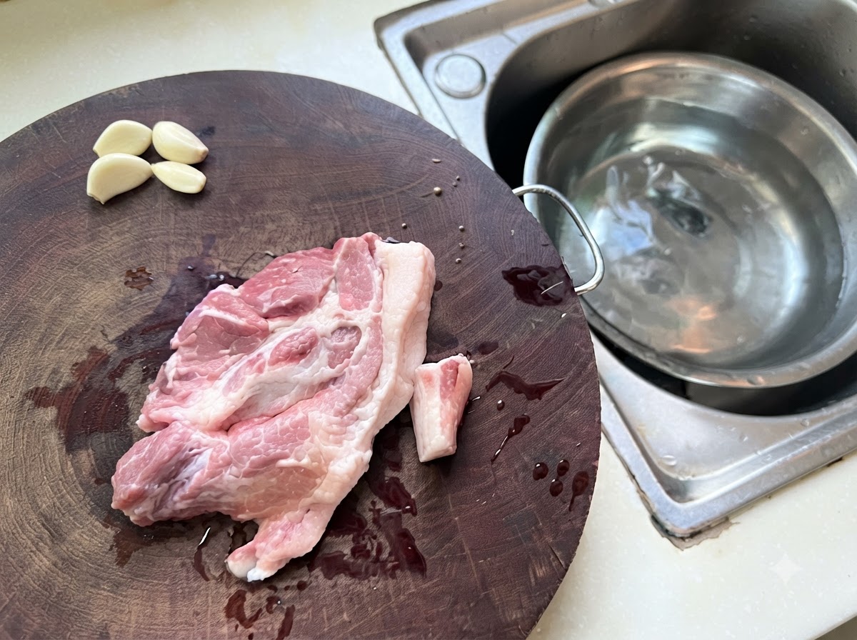 Morceaux de porc haché et dail répartis sur une planche à découper en bois à côté dun bol en métal contenant de leau.