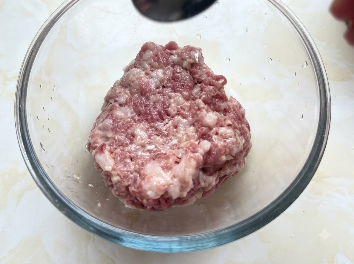 Un bol en verre rempli de porc haché recevant des assaisonnements dune cuillère tenue au-dessus.
