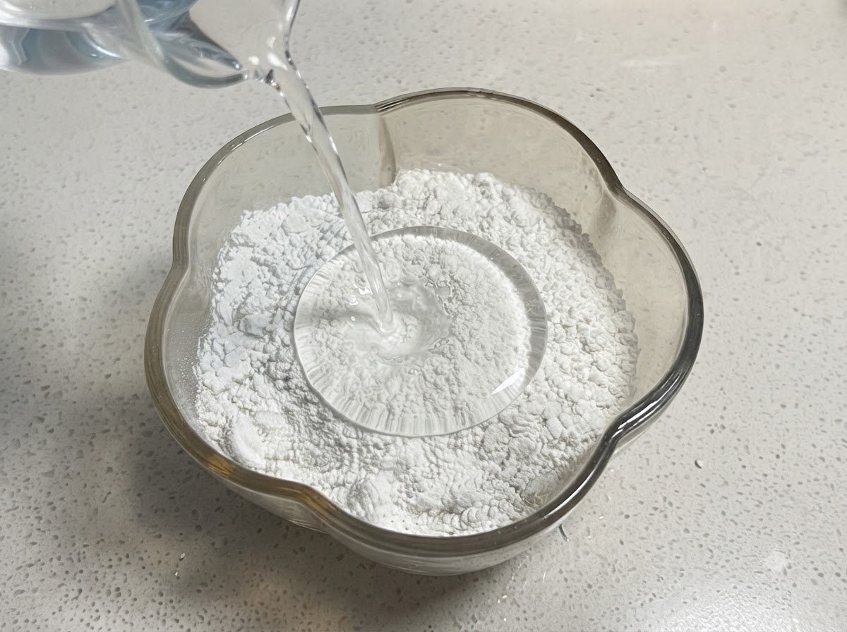 De leau versée dune tasse à mesurer dans un bol en verre contenant un mélange de farine et damidon.