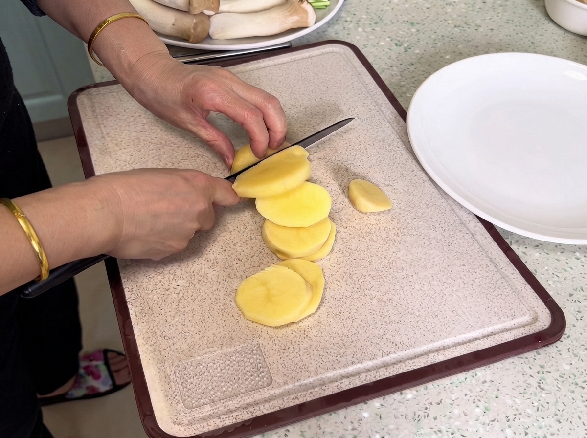 Des pommes de terre coupées avec un couteau sur une planche à découper à côté dune assiette blanche.
