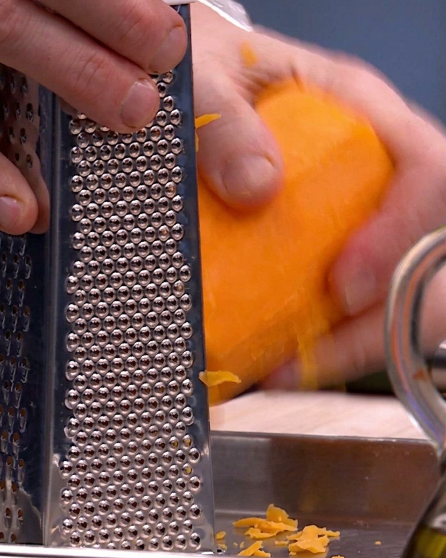 Des mains utilisant une râpe à quatre faces en métal pour râper de la courge butternut orange vif sur un plateau en métal.