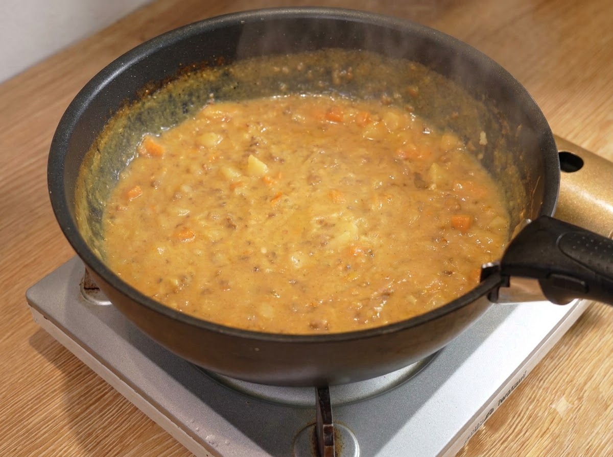 Une cuillère en bois remuant une sauce de viande au curry épaisse et bouillonnante à lintérieur dune poêle noire sur un brûleur.