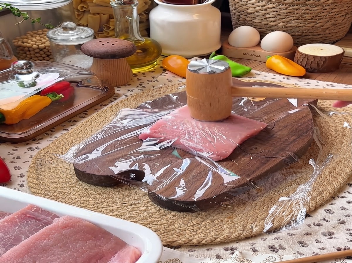 Un maillet à viande en bois écrasant un morceau de filet de porc recouverte de film plastique sur une planche à découper en bois.