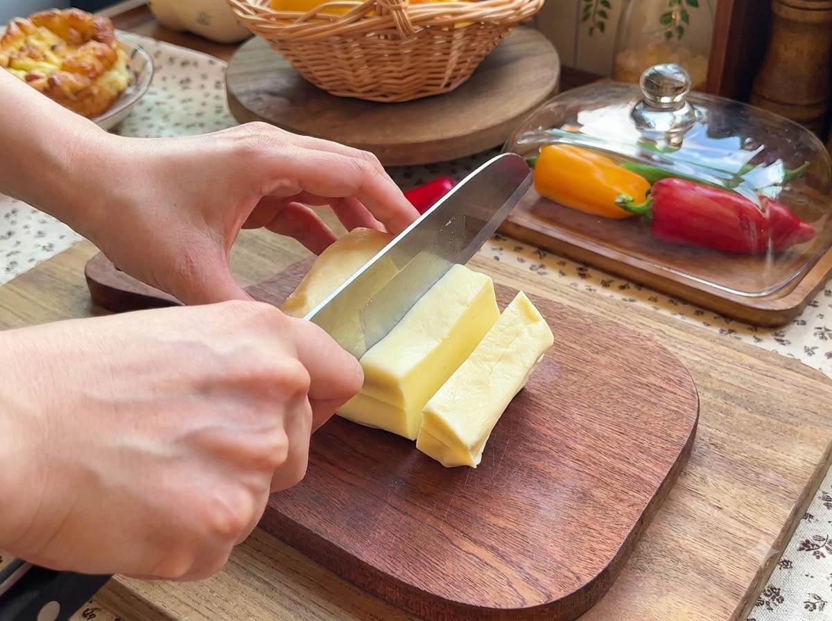 Une paire de mains utilisant un couteau pour trancher un bloc de fromage mozzarella en petits morceaux rectangulaires sur une planche à découper en bois.