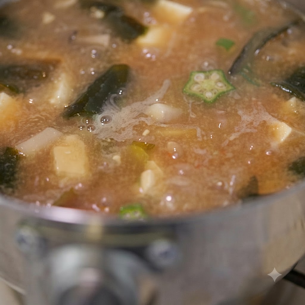 Gros plan sur la soupe miso aux gombos en train de frémir avec des morceaux visibles de gombos, de tofu et de champignons pendant quelle est remuée.