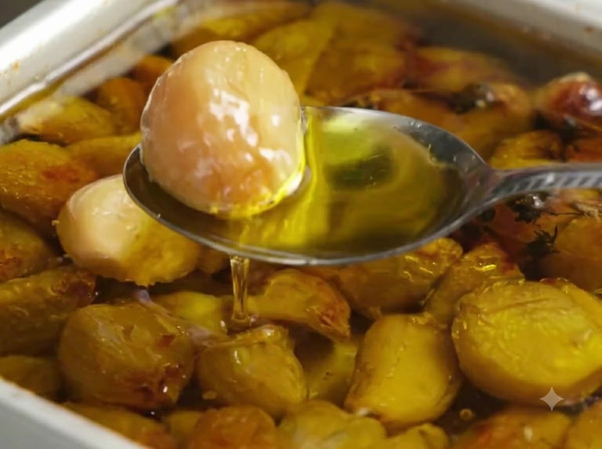 Une cuillère prélevant une gousse dail rôti dorée dun plat de cuisson rempli dhuile dolive et de gousses dail.