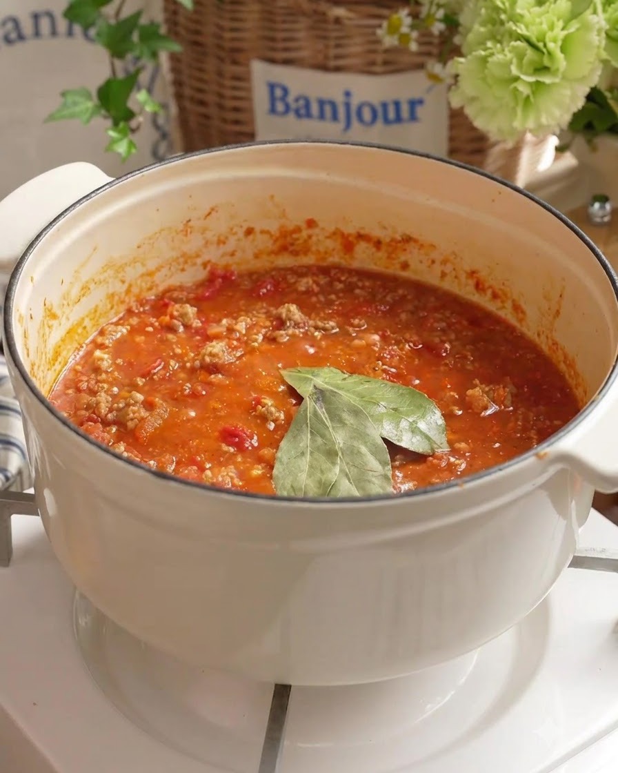 Feuilles de laurier et eau fraîche ajoutées à la sauce tomate à la viande qui mijote dans une casserole blanche.
