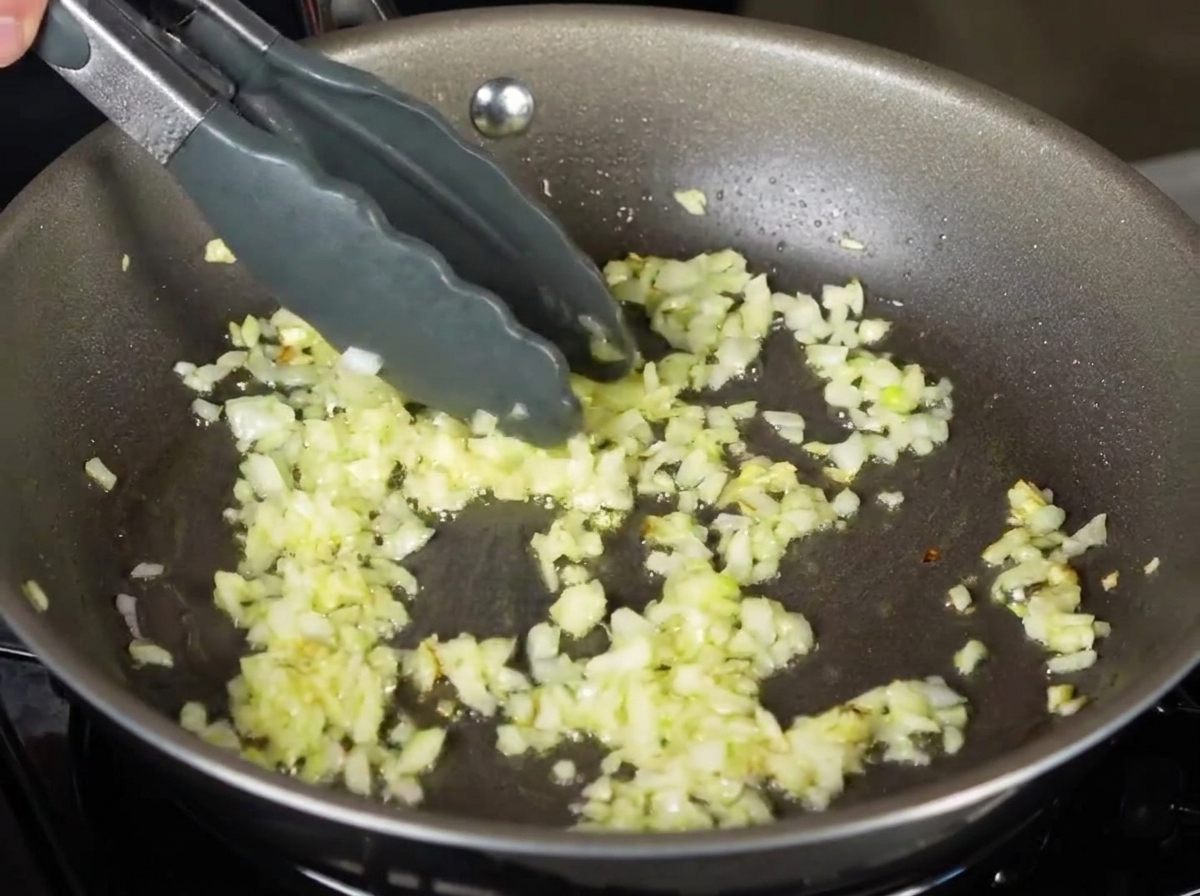 Des tagliatelles cuites placées à laide de pinces dans une poêle contenant des oignons sautés.