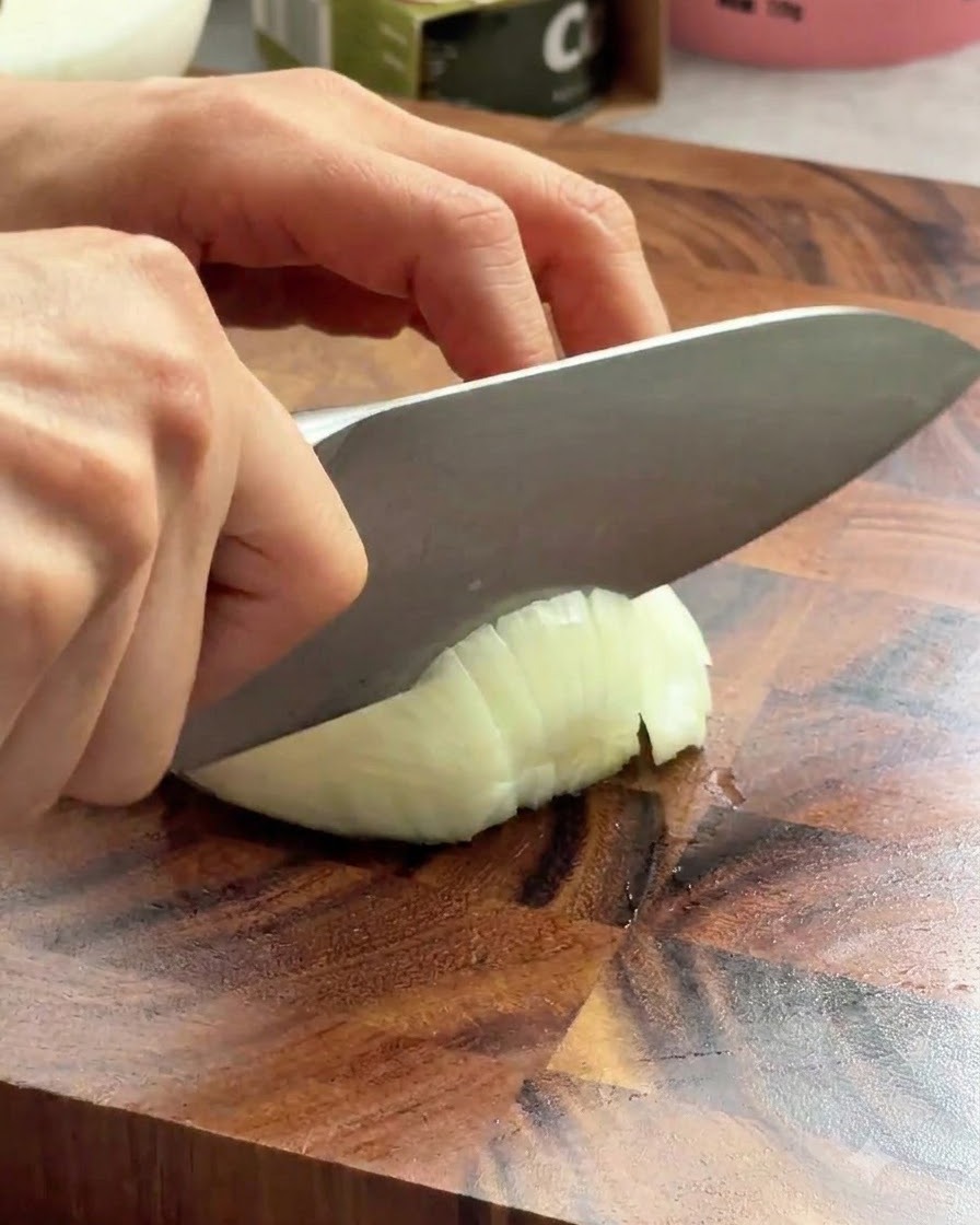 Un couteau de chef tranchant découpe un oignon blanc sur une planche en bois.