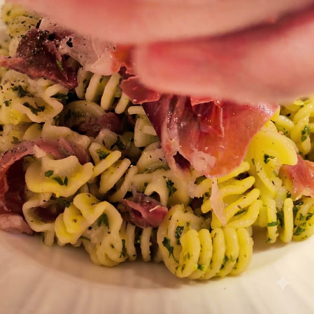 Pâtes en spirales fraîchement cuites versées dans le bol de sauce au beurre et à lail préparé, garnies dherbes fraîches et de jambon.