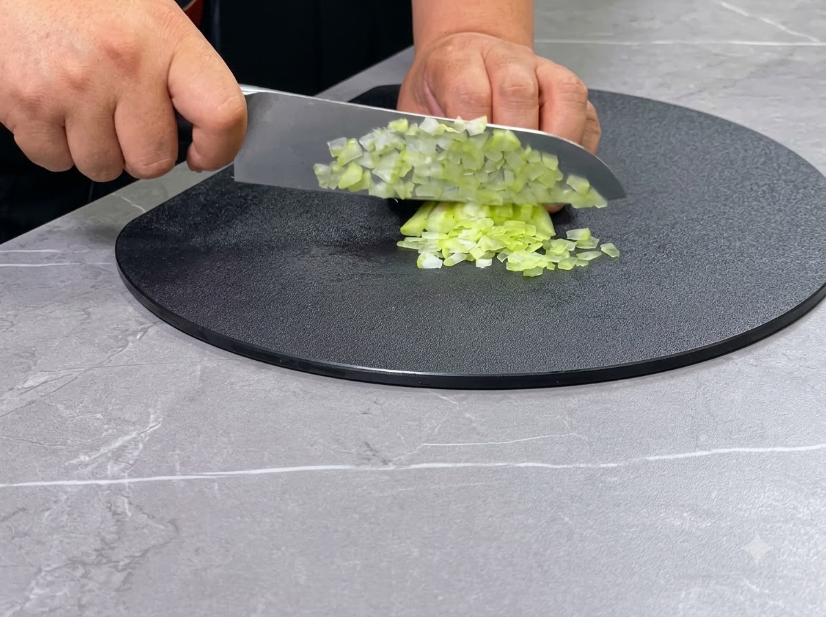 Une paire de mains utilisant un couteau de chef pour couper des branches de céleri vert en dés sur une planche à découper circulaire noire.