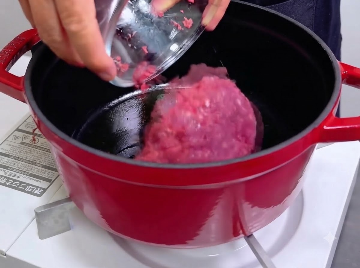 Un bol versant de la viande hachée crue et rose dans une cocotte en fonte rouge chaude.
