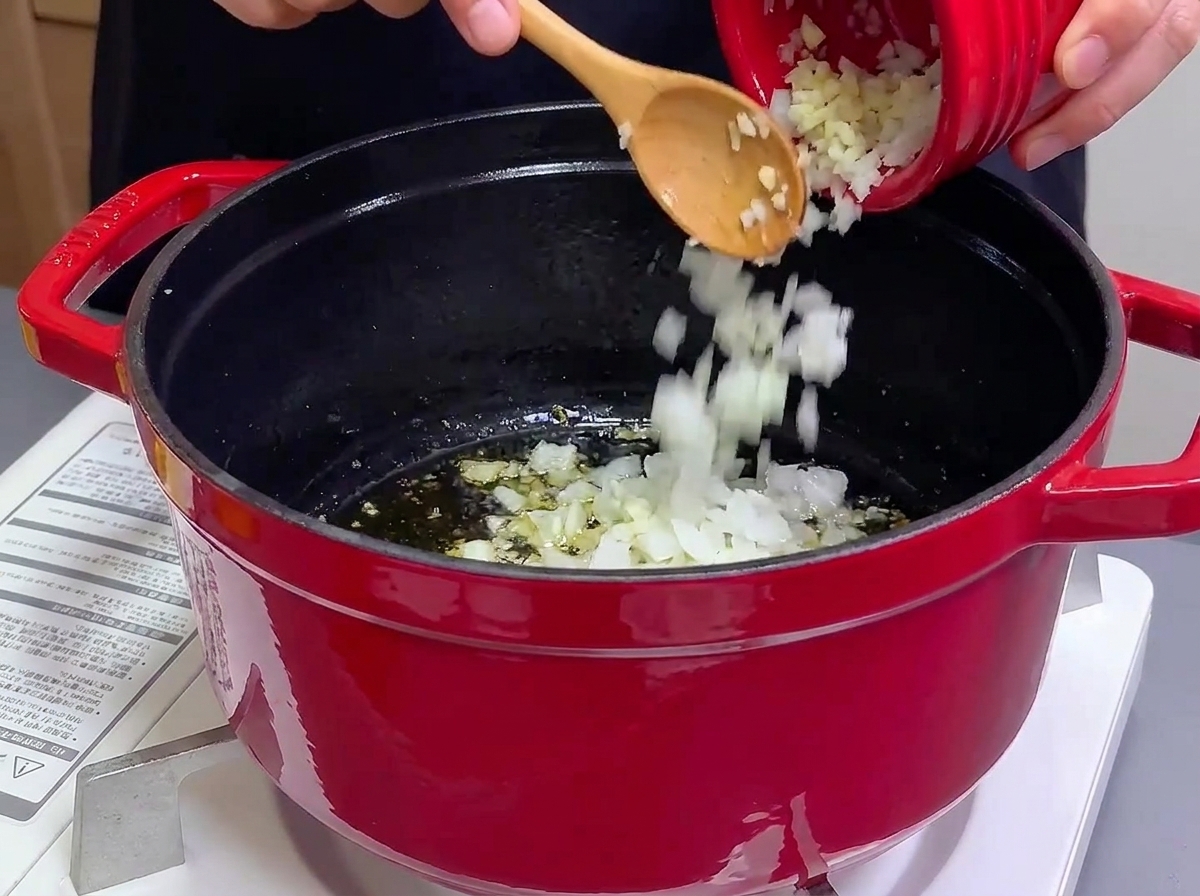 Versement doignons et dail hachés dun petit bol rouge dans une cocotte en fonte avec de lhuile chaude.