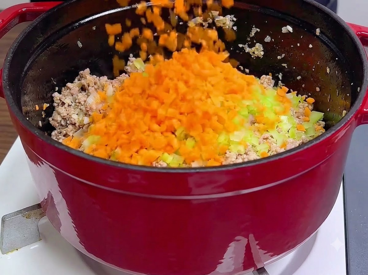 Des carottes orange et du céleri vert fraîchement hachés versés directement sur de la viande hachée cuite à lintérieur dune cocotte rouge.
