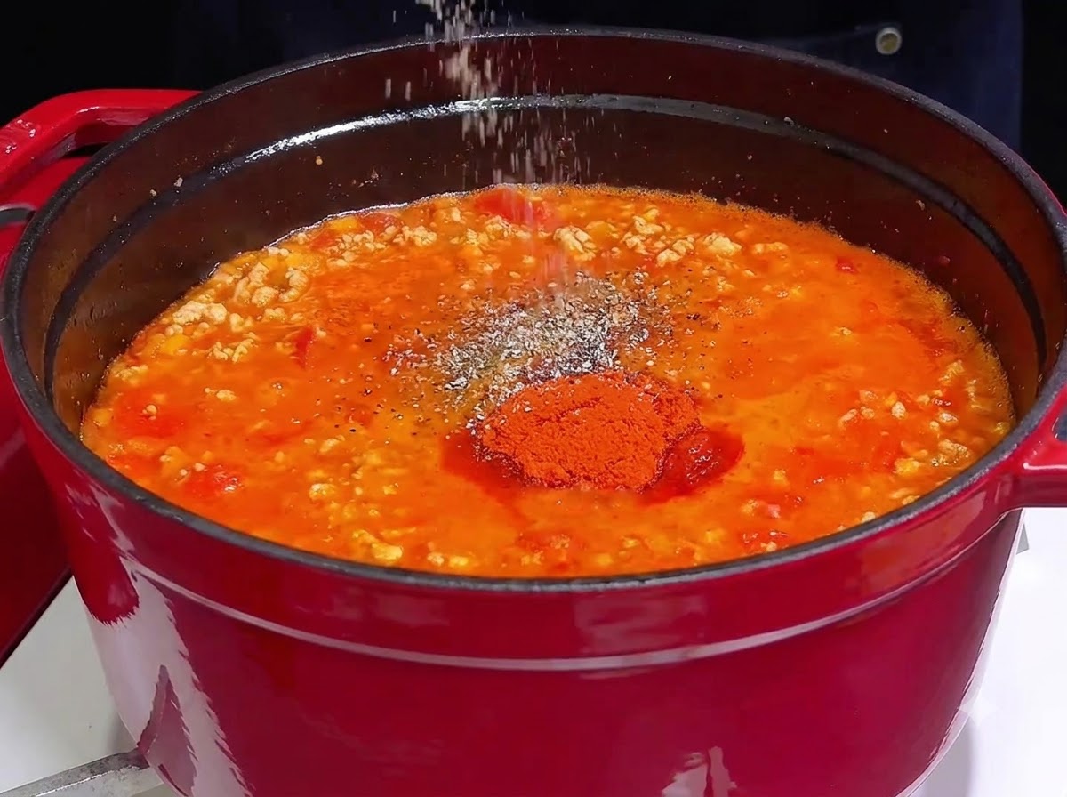 Une main saupoudrant du sel et des épices sur une sauce à la viande rouge et frémissante dans une cocotte en fonte.
