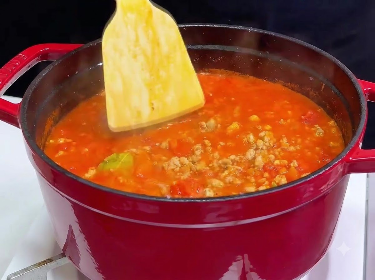 Une spatule en bois incorporant du vin rouge dans une cocotte en fonte rouge remplie de sauce tomate à la viande bouillonnante.