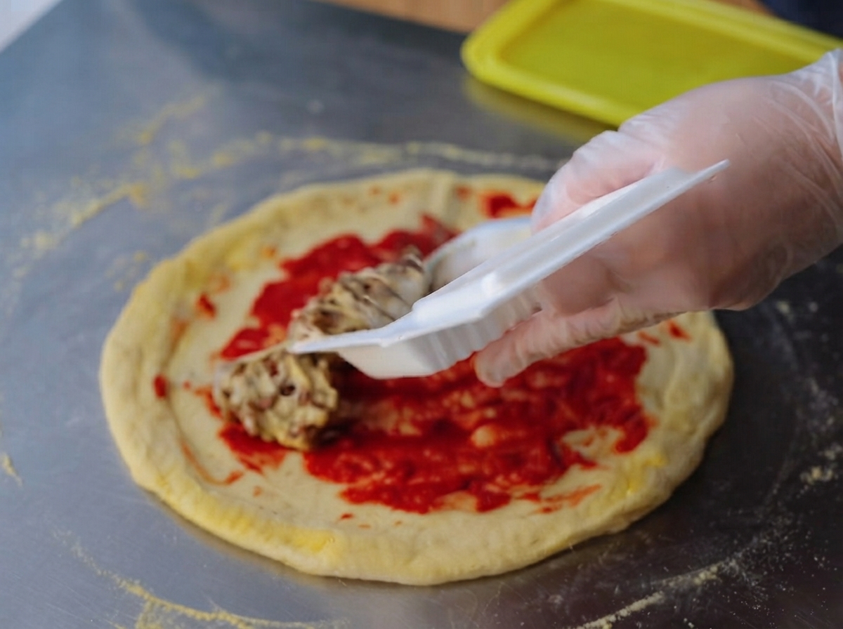 Une main gantée étalant un mélange de natto collant et filandreux uniformément sur la couche de concentré de tomate rouge vif sur une pâte à pizza crue.