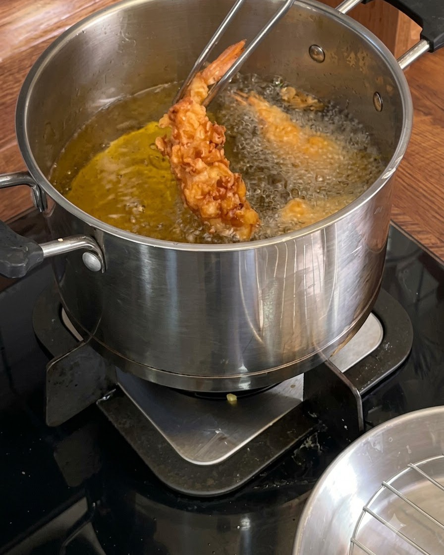 Des pinces en métal tenant une crevette frite croustillante dorée au-dessus dune casserole dhuile chaude.
