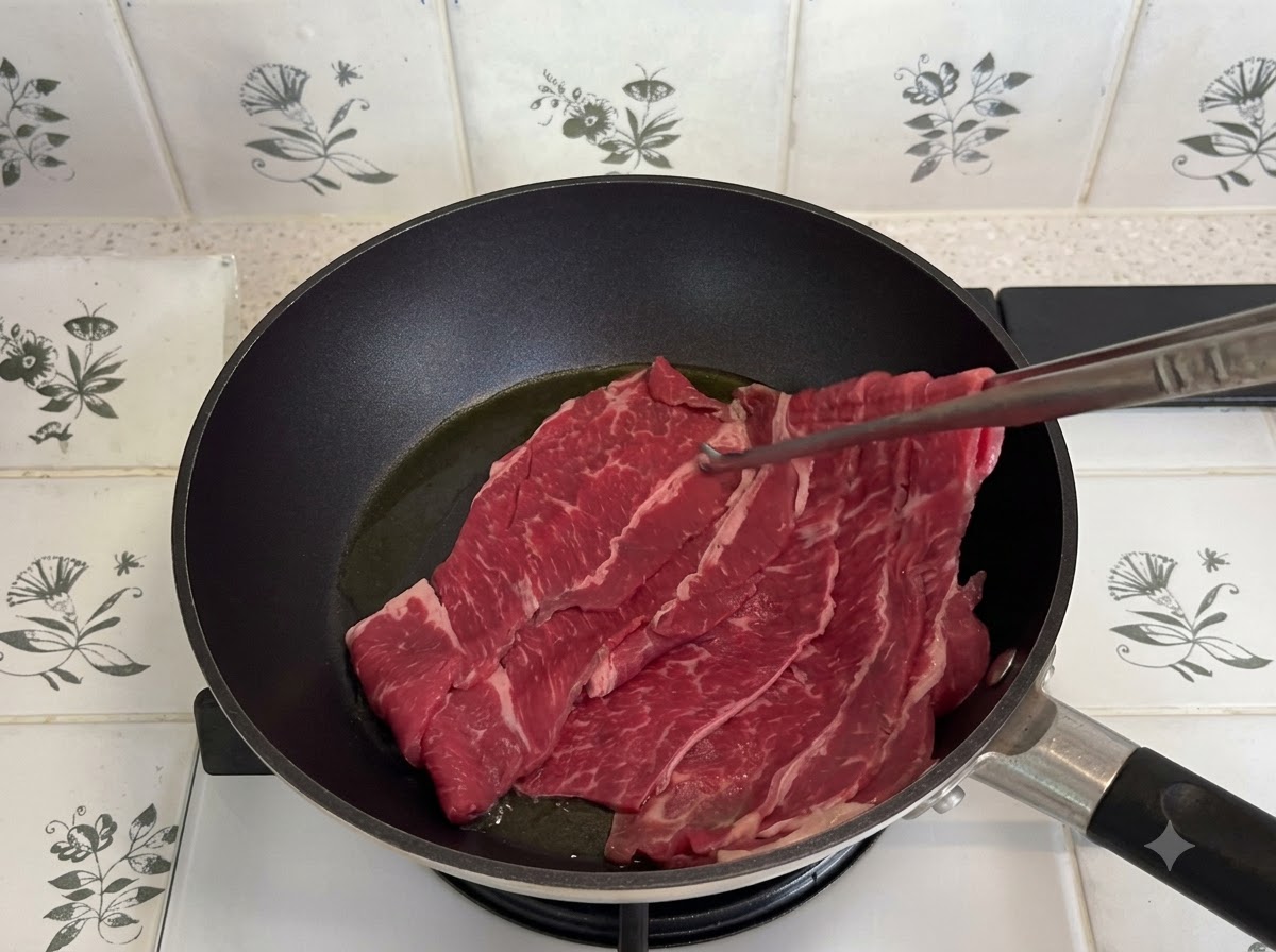 Fines tranches de bœuf gras en train de cuire dans une poêle sombre à laide de pinces de cuisine.