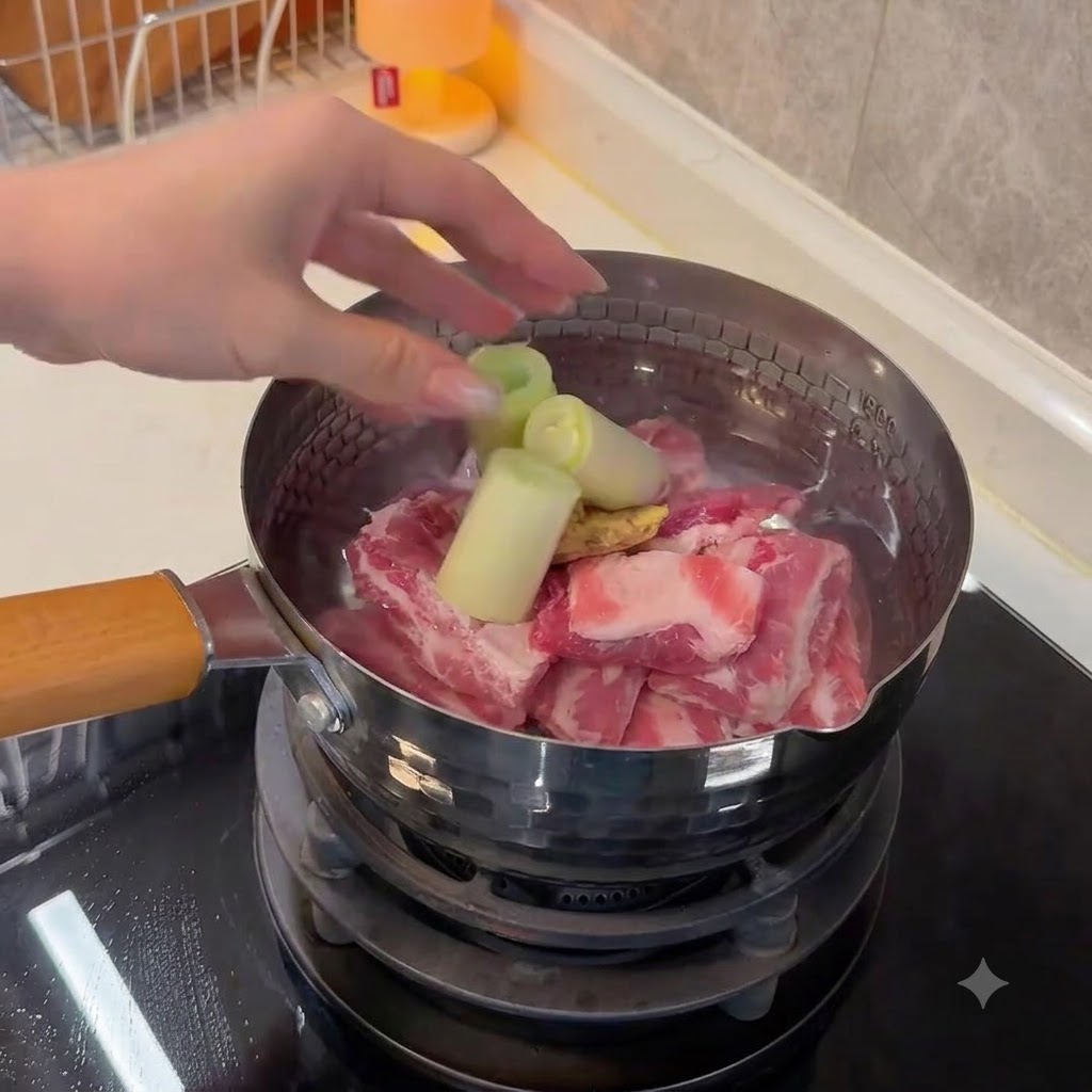 Une main ajoutant de gros tronçons de cébette et des tranches de gingembre dans une casserole deau avec le porc.