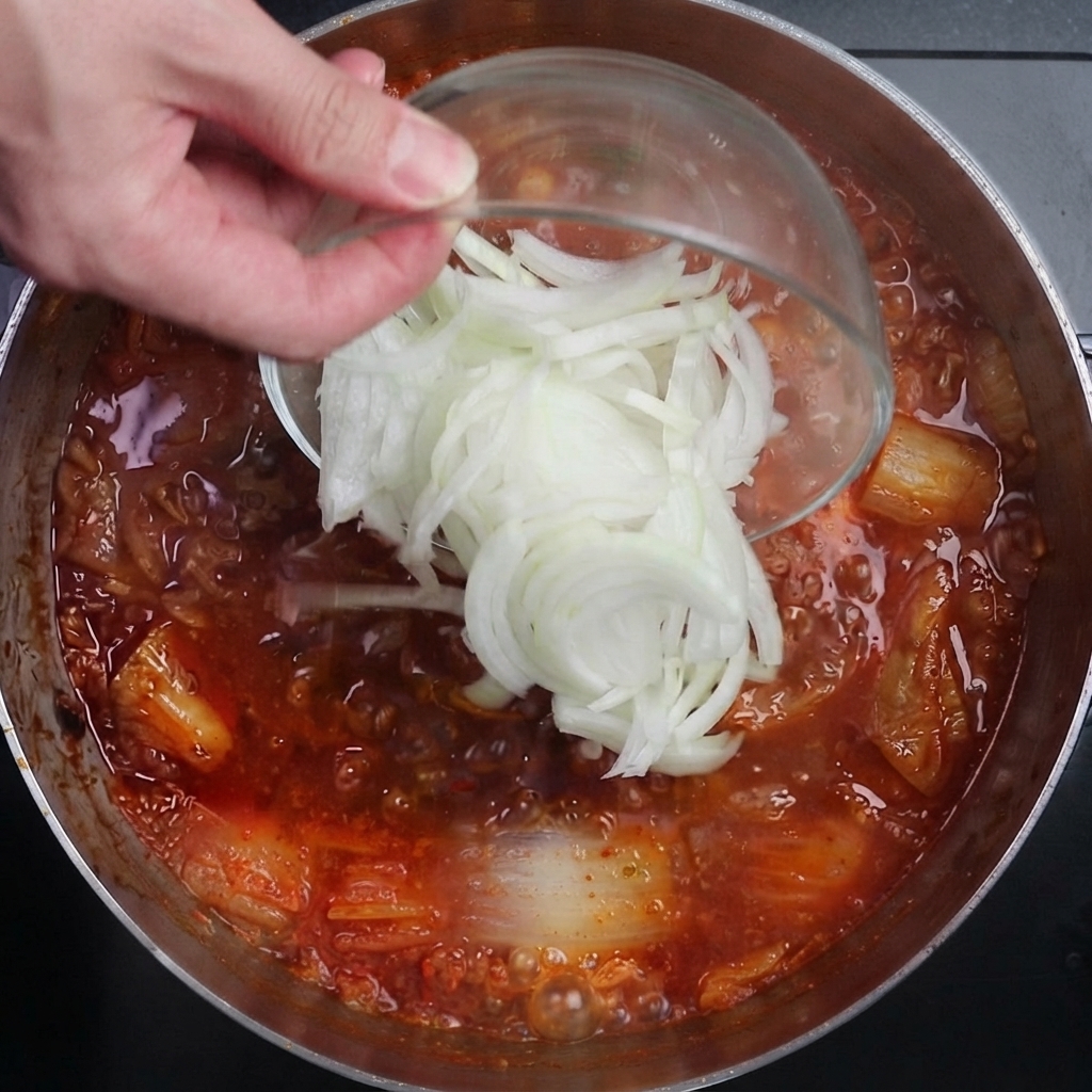 Des tranches doignons blancs versées dun bol en verre dans une casserole de bouillon de kimchi rouge épicé qui bouillonne vigoureusement.