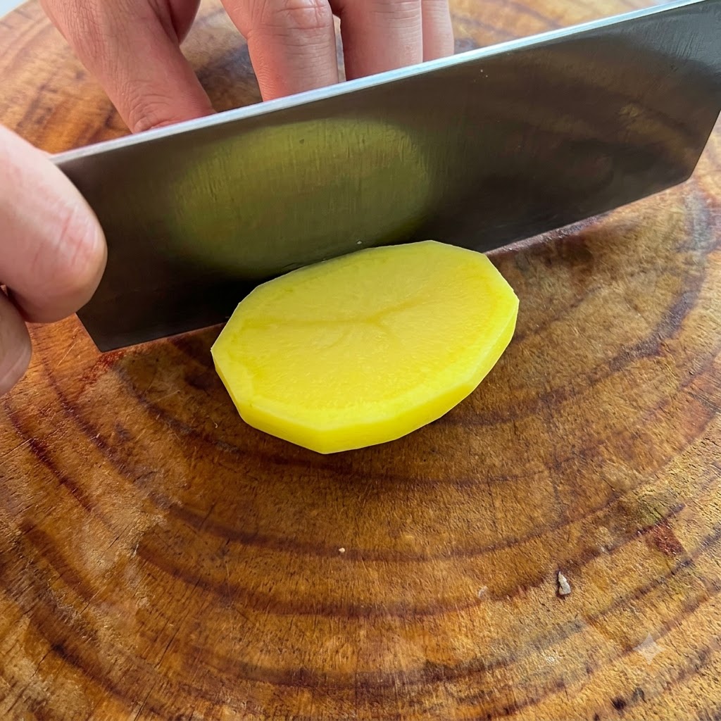 Un couteau coupant une pomme de terre jaune épluchée en tranches épaisses sur une planche à découper en bois.