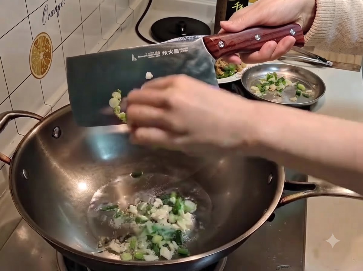 Une main utilisant un grand couperet en métal pour pousser des cébettes et de lail hachés dans lhuile chaude dun wok.