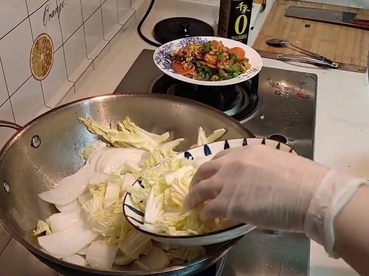 Une main gantée transférant du chou napa cru fraîchement coupé dune assiette à motifs dans un wok.