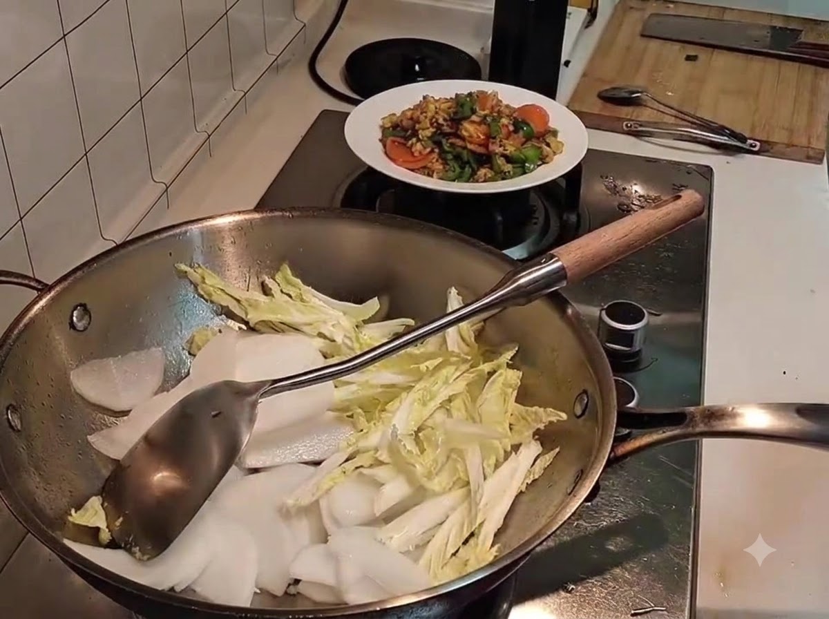 Des tranches de radis blanc reposant sur du chou napa légèrement flétri à lintérieur dun wok en métal, avec une spatule à proximité.