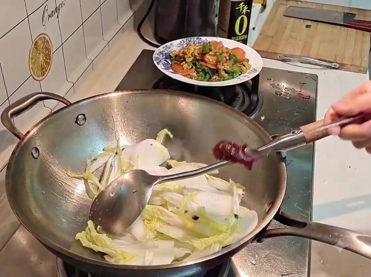 Une main tenant une cuillère en métal chargée de pâte de piment rouge foncée au-dessus dun wok rempli de chou et de radis.