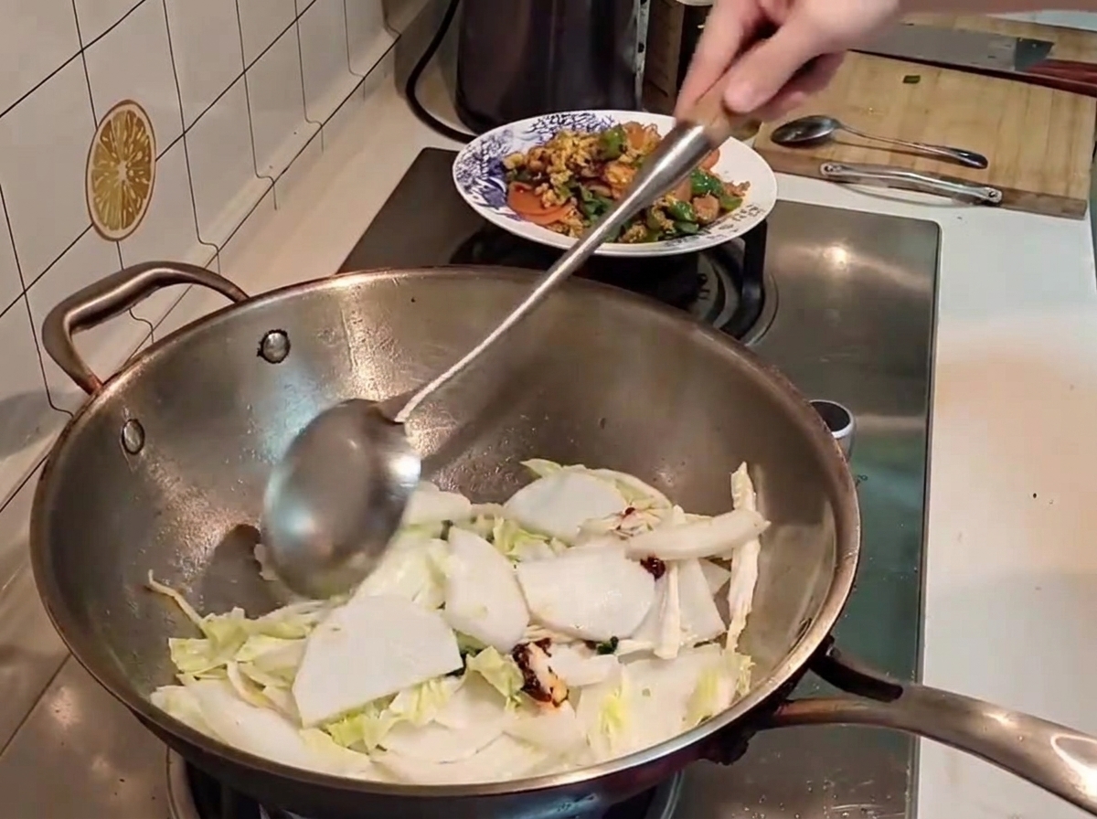 Une spatule en métal mélangeant des morceaux de chou napa et de radis blanc avec de la pâte de piment rouge dans un wok très chaud.