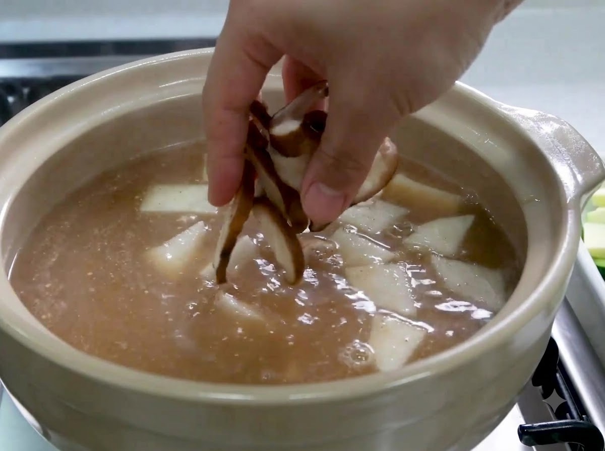 Une main ajoutant des champignons shiitake émincés dans la marmite contenant le bouillon et le radis.