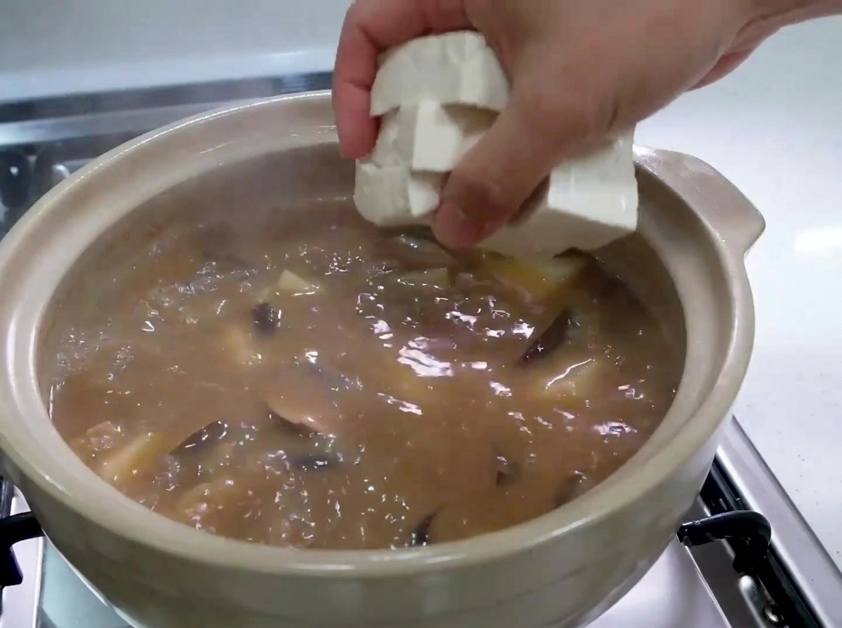 Une main déposant délicatement des cubes de tofu blanc frais dans un ragoût bouillonnant.