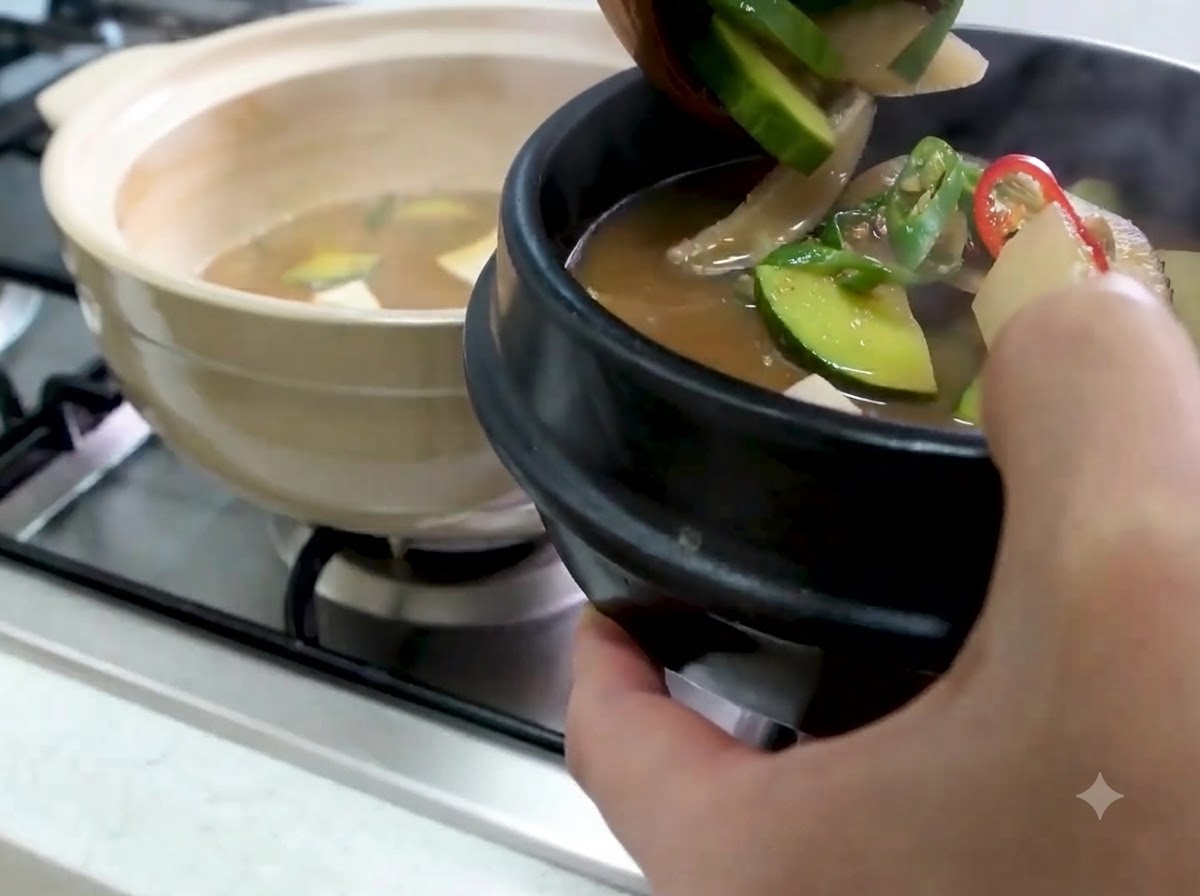 Une louche transférant le Doenjang Jjigae chaud avec courgette et légumes dans un bol de service en pierre noire.