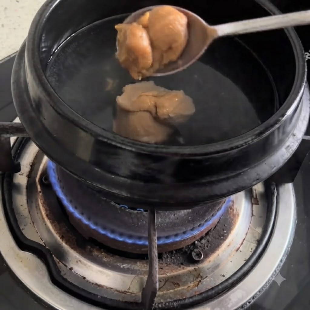 Une cuillerée de pâte de soja versée dans une casserole noire deau bouillante sur une cuisinière à gaz.