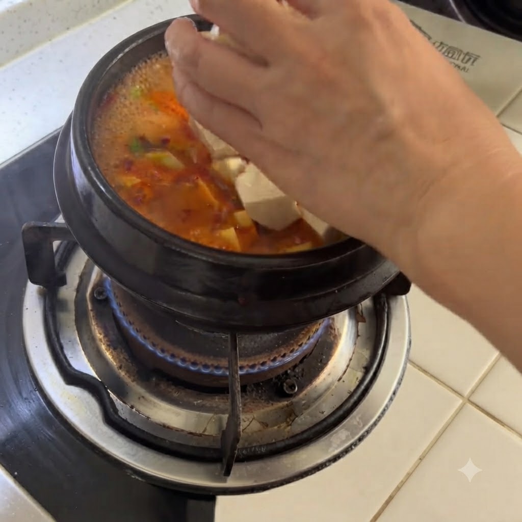 Des cubes de tofu blanc déposés à la main dans un pot en argile frémissant de soupe épicée.
