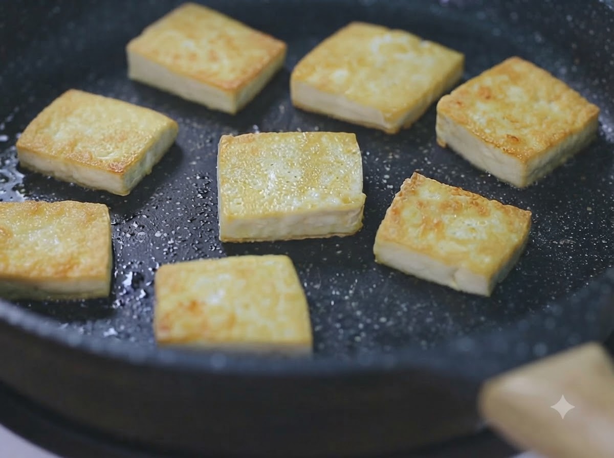 Plusieurs cubes de tofu dorés grésillant dans une poêle noire à feu doux.